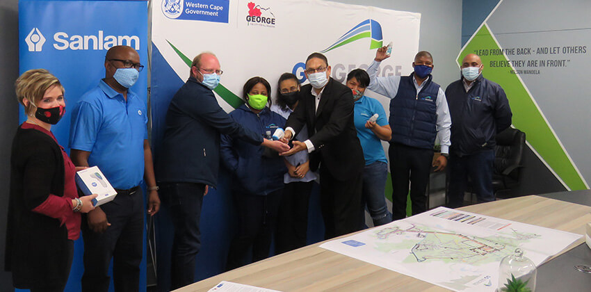 Man testing the non-contact infrared thermometer on another man’s hand while a number of people are looking on. Everybody is wearing face masks covering their mouths and noses.