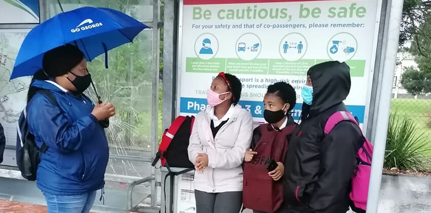 Three school children standing under a bus shelter while a woman in GO GEORGE uniform talks to them. It is raining and the woman is using an umbrella.