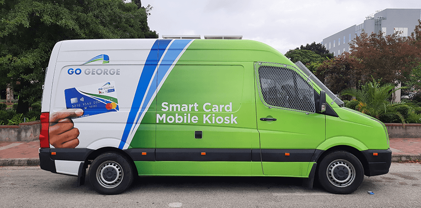 A minibus branded in the GO GEORGE colours of white, green and blue with a picture of a GO GEORGE Smart Card on the side. The writing on the minibus reads, “Smart Card Mobile Kiosk”.
