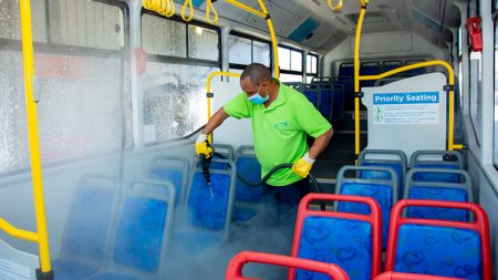 A man wearing a face mask, spraying the the seats inside a bus with a sprayer that emits a strong cloud of steam.