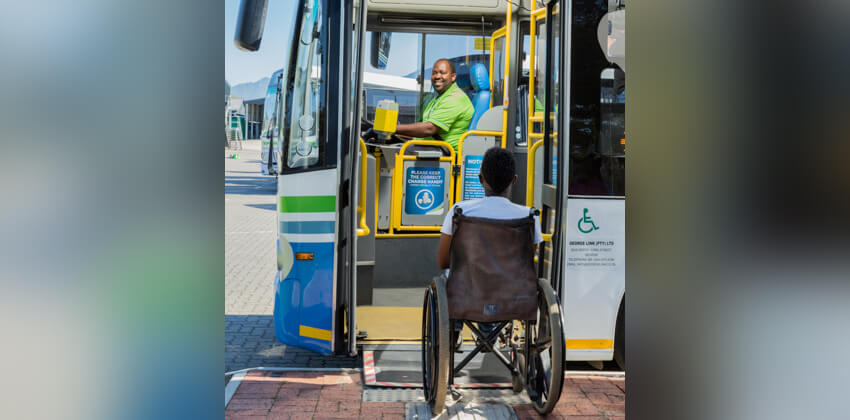 A passenger in a wheelchair boarding a large GO GEORGE bus using the deployed ramp which gives same-level access to the bus. A friendly bus driver is looking on from his driver’s seat.