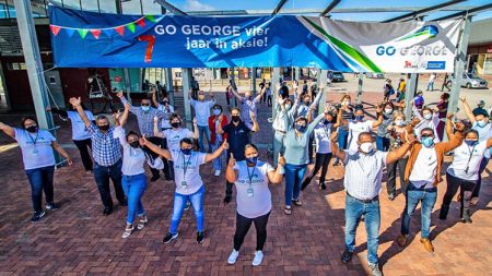 A crowd of about 30 people in an open space waving excitedly in front of a banner with the message: GO GEORGE celebrates 7 years in action!