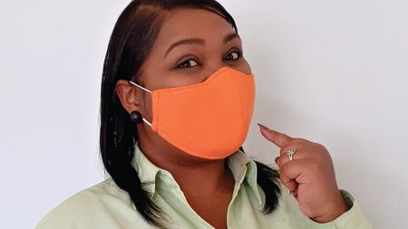 A smiling young woman pointing to a mask that covers both her mouth and nose.