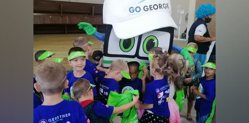 A group of toddlers flocking to greet and talk to the GO GEORGE mascot, Georgie. The mascot looks like a bus, with a friendly face and huge eyes. The little ones are not wearing face masks.