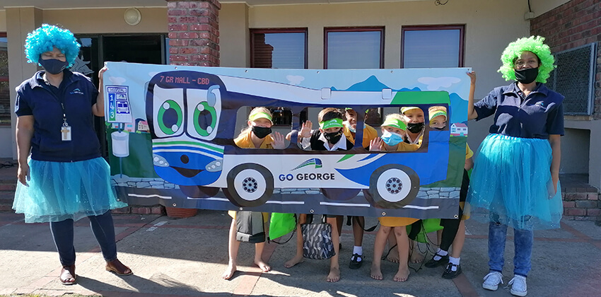 Three children of about 10 years old are peeping through the cut-out windows of a bus-shaped banner held by two women in blue and green costume and colourful wigs.