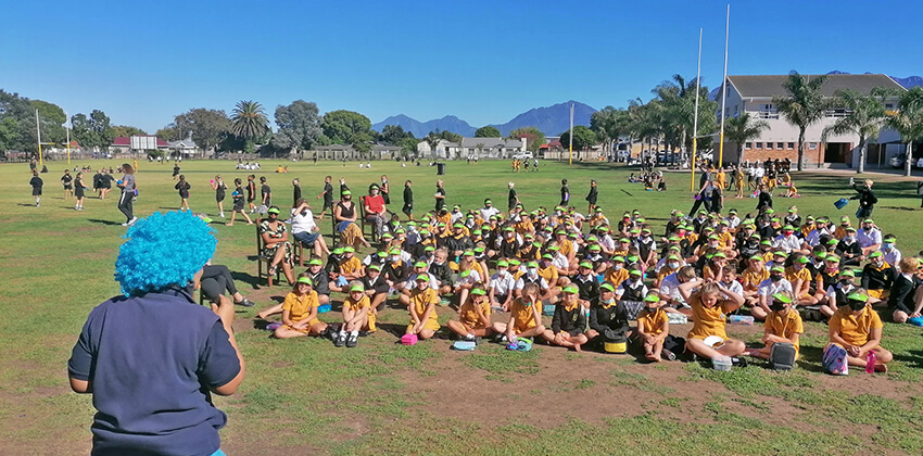 A group of grade 4 learners sitting on a sports field, addressed over a microphone by a lady in blue costume and a blue wig.  