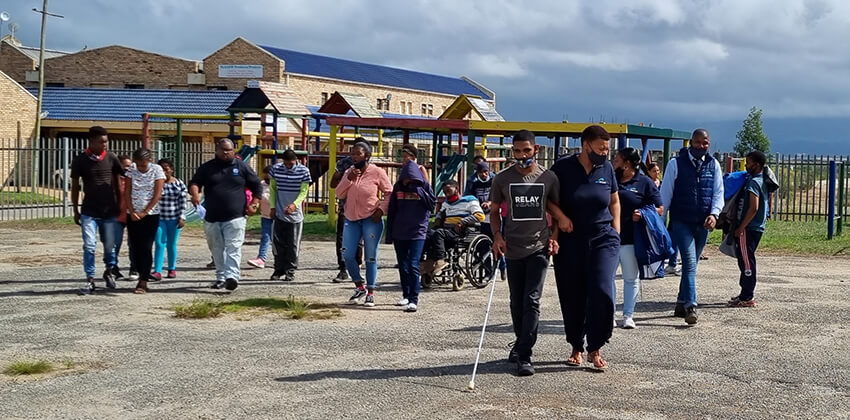 A group of people walking together. There are people using wheelchairs and a blind person is walking arm in arm with a lady wearing a GO GEORGE uniform.