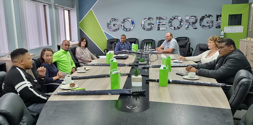 A group of people in discussion around a boardroom table with the name GO GEORGE on the wall behind them.