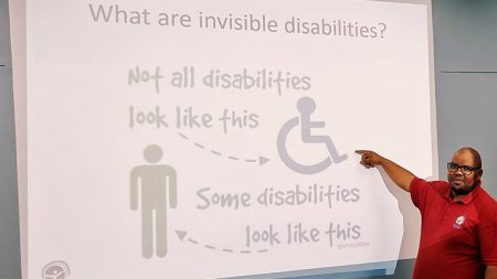 A man standing at a digital board, pointing to a slide that describes the fact that disabilities may be visible or invisible.