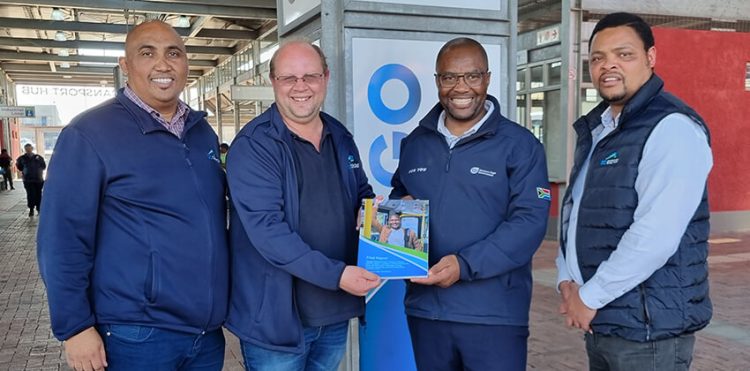 Four men standing in front of a GO GEORGE-branded background. Two of them are holding onto a booklet.