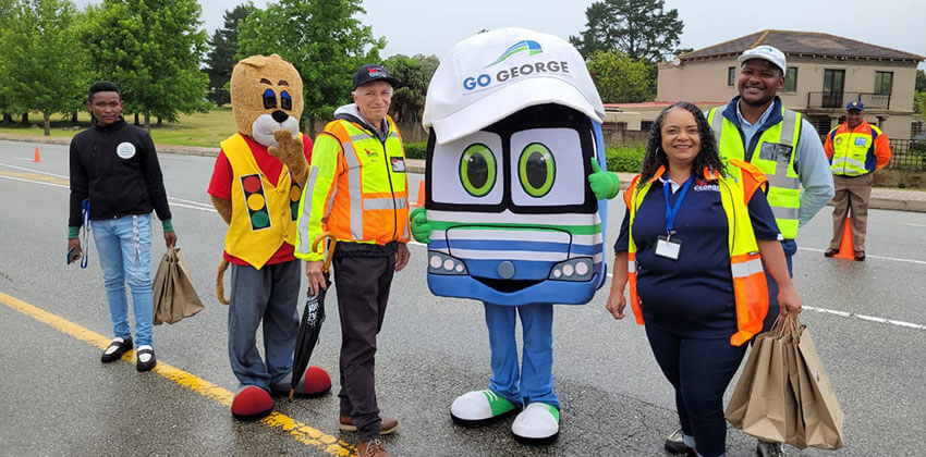 People in reflective jackets and umbrellas along with two human-size mascots standing in the road, welcoming motorists entering George for the holiday.