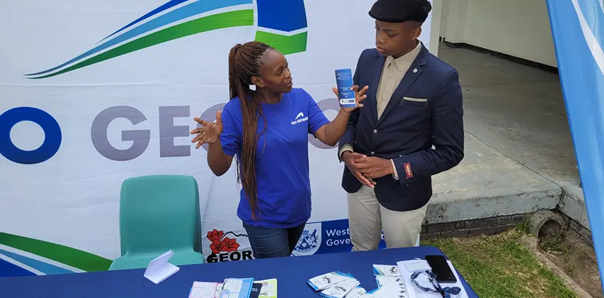 A woman in a GO GEORGE uniform and a young man talking in front of a huge screen with the GO GEORGE logo on. In front of them is a table with a display of pamphlets.