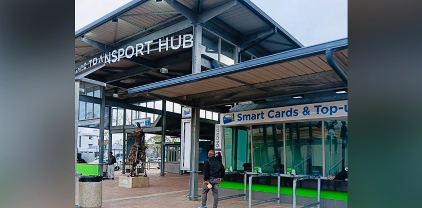 Exterior view of the revamped GO GEORGE Transport Hub, featuring a modern steel-and-glass canopy structure with "TRANSPORT HUB" signage. A green-and-white kiosk on the right displays a "Smart Cards & Top-Up" sign for ticketing services. A bronze statue stands near the entrance, with a pedestrian walking toward the building and another person visible in the background under the covered walkway.