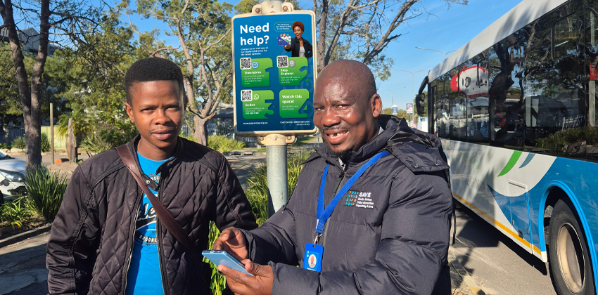 A GO GEORGE surveyor wearing a branded lanyard shows a smartphone screen to a young passenger at a bus stop, with a GO GEORGE bus and an information signpost in the background.